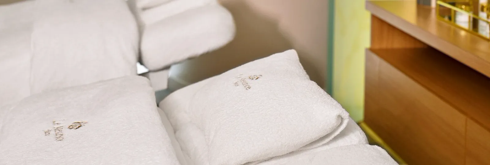Spa room with a reclining massage chair covered in white towels. The soft glow of ceiling lights creates a serene ambiance. A bouquet of flowers sits on a wooden counter, adding elegance.