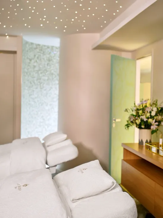 Spa room with a reclining massage chair covered in white towels. The soft glow of ceiling lights creates a serene ambiance. A bouquet of flowers sits on a wooden counter, adding elegance.