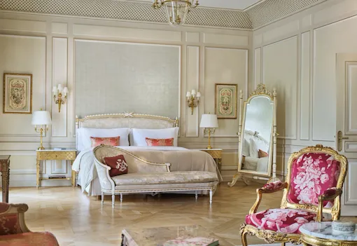 Chambre élégante à la décoration vintage. Elle dispose d'un lit luxueux, d'un miroir orné d'un cadre doré, de chaises fleuries et d'un éclairage tamisé, évoquant une atmosphère classique et opulente.