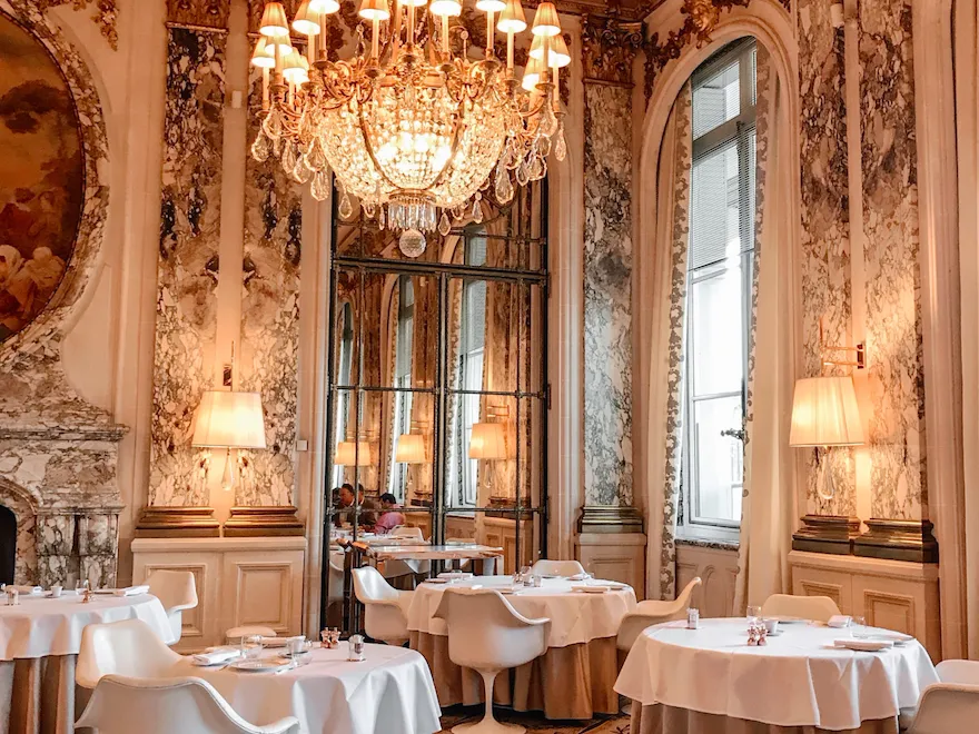 Salle de restaurant avec un grand lustre éclairant les ornements dorés sur les murs. Au Meurice, Paris