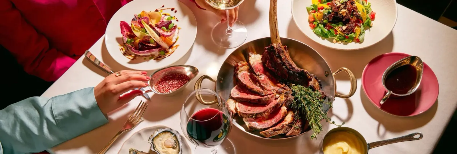 Dining table at The Grill at The Dorchester, showcasing a sliced Tomahawk steak served on the bone, accompanied by oysters, seasonal salads, rich sauces and glasses of red wine in an elegant sharing-style feast.