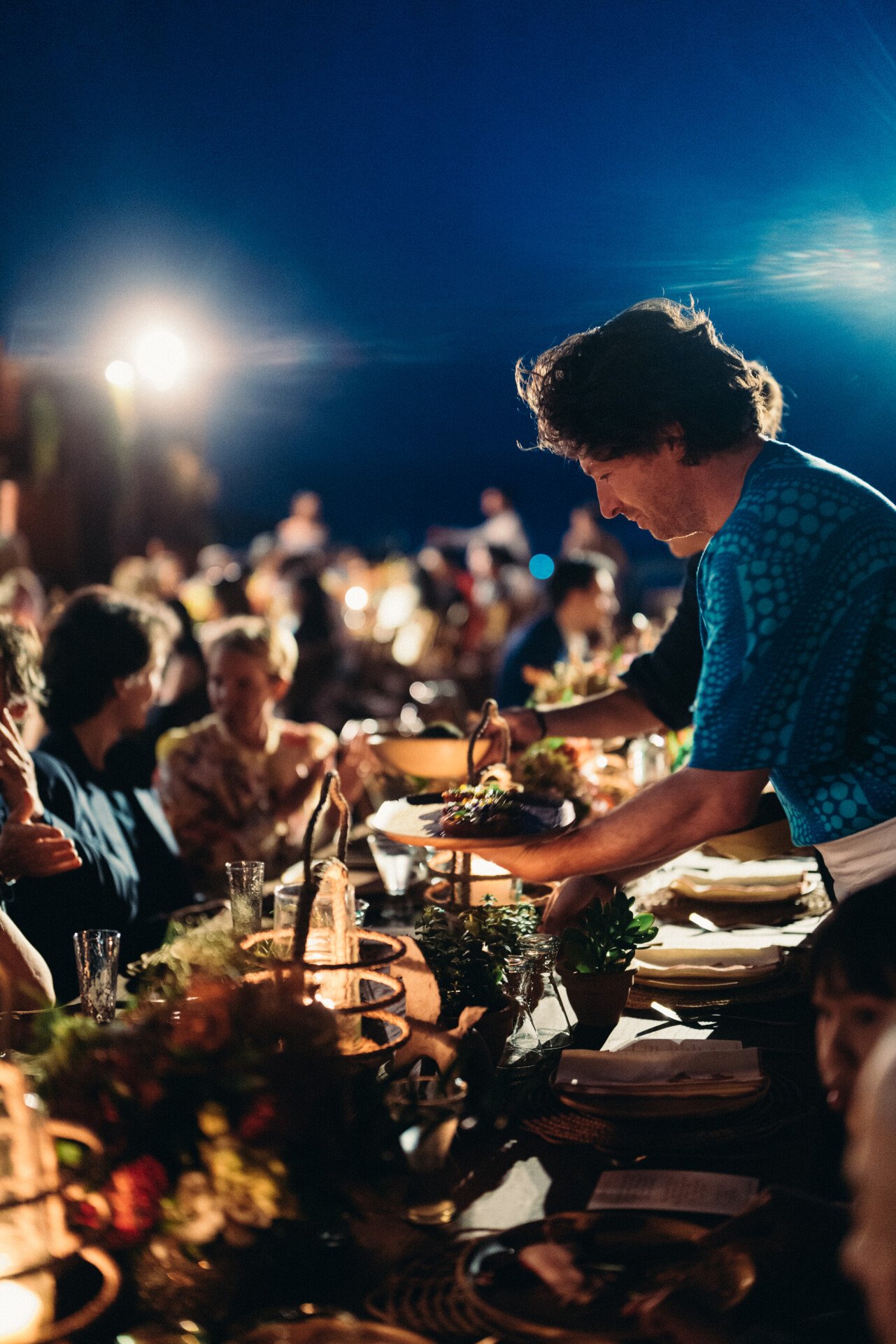 Une personne vêtue d'une chemise bleue sert le repas lors d'un dîner festif à l'éclairage chaleureux. Les convives sourient et discutent autour d'une table décorée de plantes et de bougies.