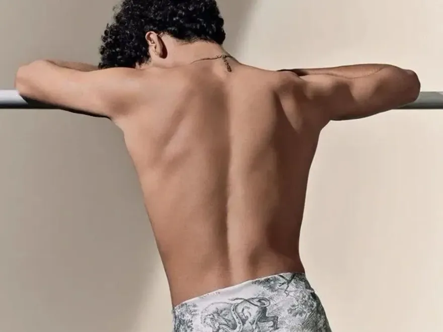 A shirtless man leans against a bar with his elbow resting on it, showcasing his muscular shoulder and chest. He is indoors, and the backdrop includes a wall visible behind him.