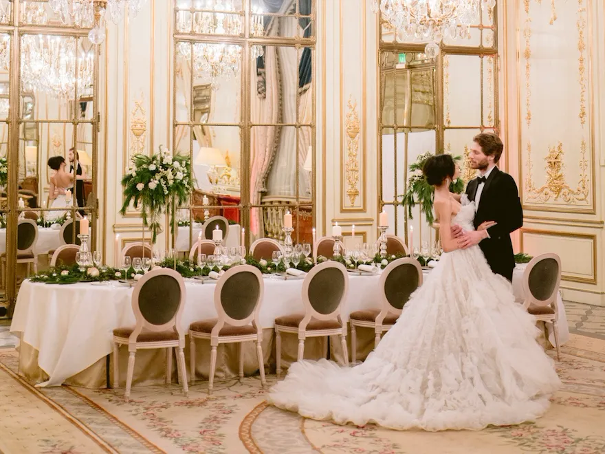 Wedding couple standing in front of a festive set table in Salon Pompadour. Green and white flower arrangements are on the table. The bride is looking at her husband, at Le Meurice, Paris