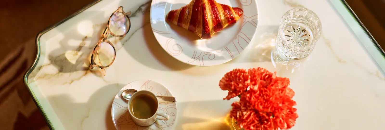 White marble dining table at la galerie seen from above, with reading glasses, a cup of coffee, a croissant on a plate and a red geranium, at Hotel Plaza Athénée, Paris