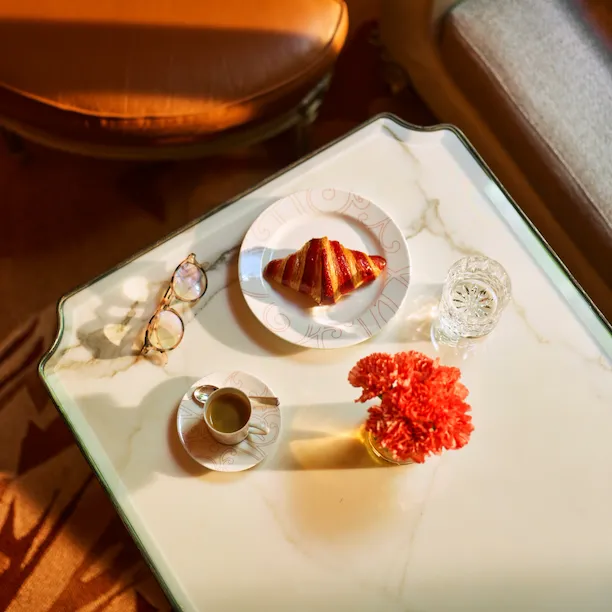 White marble dining table at la galerie seen from above, with reading glasses, a cup of coffee, a croissant on a plate and a red geranium, at Hotel Plaza Athénée, Paris