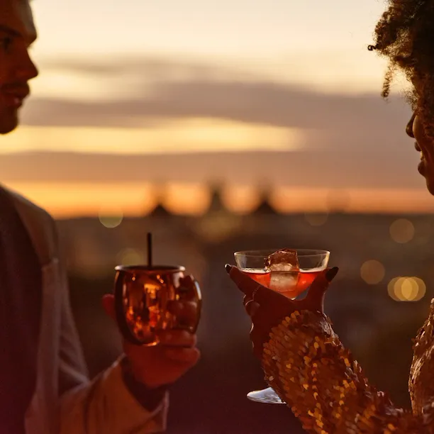 Un giovane uomo e una giovane donna con due drink che parlano con lo sfondo di un tramonto su Roma
