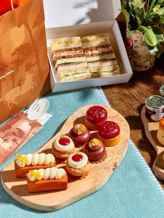 Takeaway meadow afternoon tea with finger sandwiches in box, summer pastries, scones, cream and jam and champagne layed out on picnic blanket with takeaway bag for the Royal Windsor Flower Show
