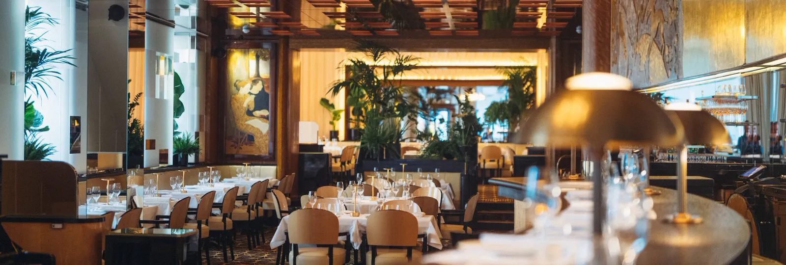 Intérieur élégant avec une ambiance chaleureuse et accueillante. Les tables sont dressées avec des nappes blanches impeccables, entourées de chaises beiges. Des plantes vertes et un éclairage tamisé renforcent l'atmosphère cosy.