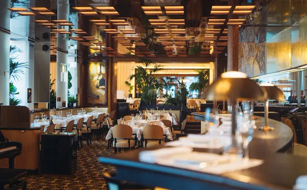 Intérieur élégant avec une ambiance chaleureuse et accueillante. Les tables sont dressées avec des nappes blanches impeccables, entourées de chaises beiges. Des plantes vertes et un éclairage tamisé renforcent l'atmosphère cosy.