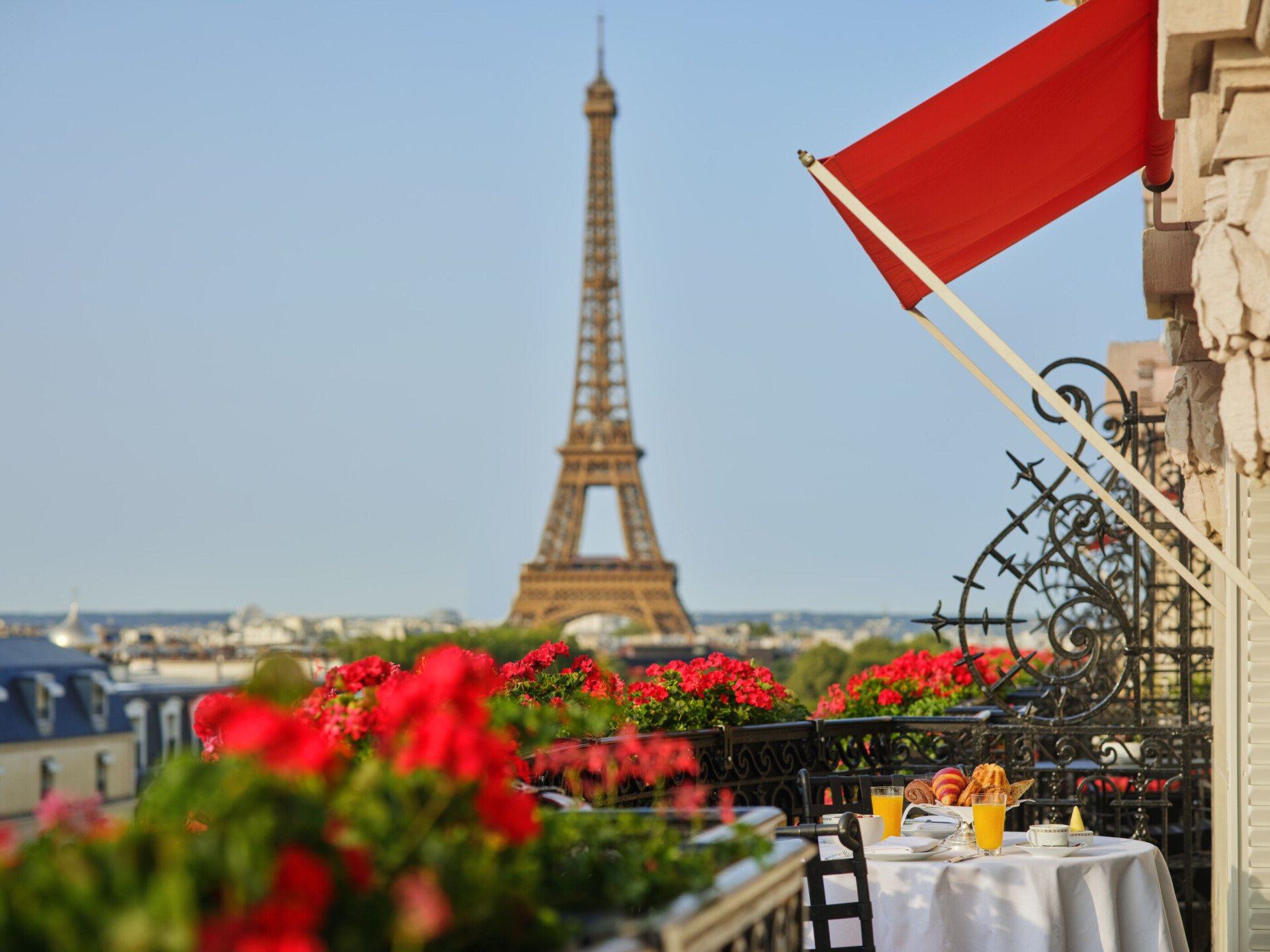 Vista da Torre Eiffel do terraço, com flores vermelhas vibrantes em primeiro plano. Uma mesa posta para o pequeno-almoço com croissants e sumo de laranja transmite uma manhã serena em Paris.