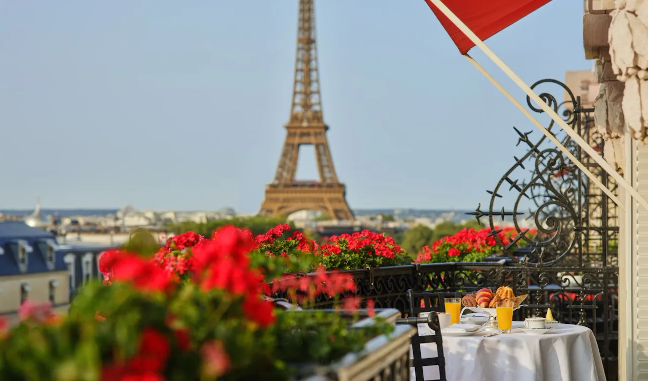 Vista da Torre Eiffel do terraço, com flores vermelhas vibrantes em primeiro plano. Uma mesa posta para o pequeno-almoço com croissants e sumo de laranja transmite uma manhã serena em Paris.