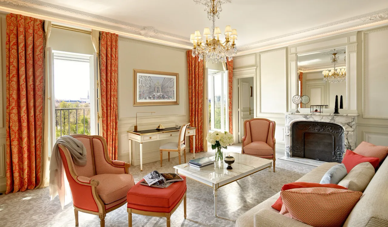 Salon luxueux avec lustre orné, rideaux rouge-orange et mobilier élégant autour d'une cheminée en marbre. Ambiance lumineuse et sophistiquée.