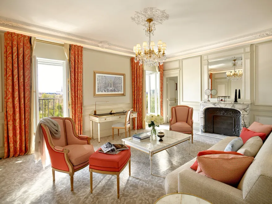 Salon luxueux avec lustre orné, rideaux rouge-orange et mobilier élégant autour d'une cheminée en marbre. Ambiance lumineuse et sophistiquée.