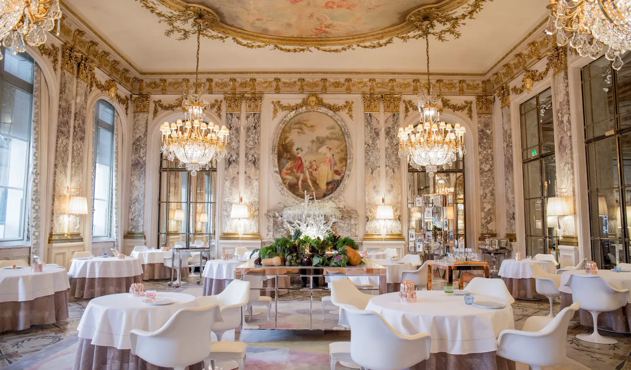 Luxurious dining room with ornate chandeliers, elegant round tables, and white chairs. Large murals and decorative gold accents create an opulent ambiance.