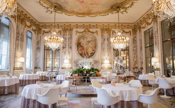Salle à manger luxueuse avec lustres décorés, tables rondes élégantes et chaises blanches. De grandes peintures murales et des accents décoratifs dorés créent une ambiance opulente.
