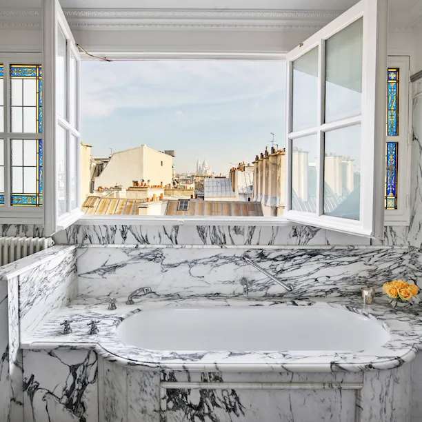 Salle de bains en marbre avec fenêtres ouvertes donnant sur les toits de Paris dans une suite junior exécutive au Meurice, Paris.