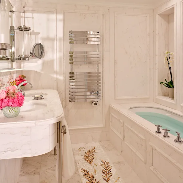 Banheiro elegante com superfícies em mármore, com pia branca decorada com flores, toalheiro cromado e banheira com decoração de orquídeas, exalando luxo.