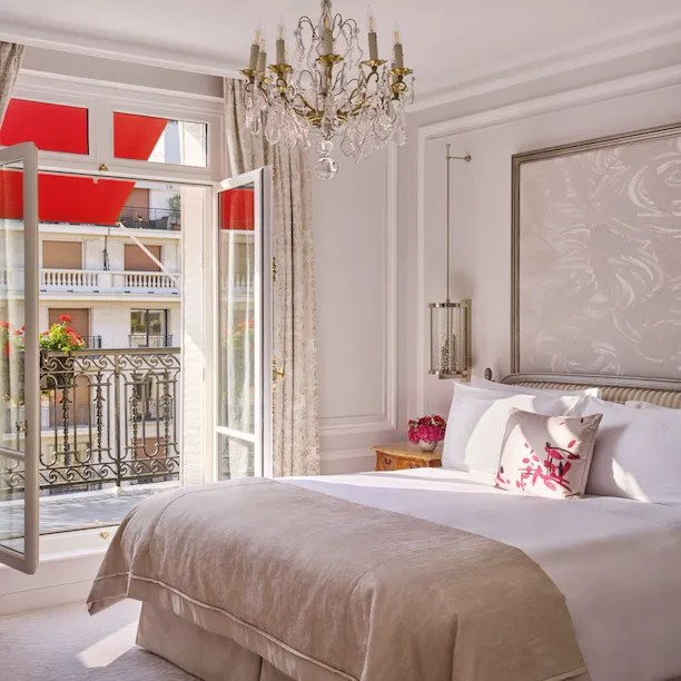 Quarto elegante com cama bege, lustre ornamentado e portas da varanda abertas revelando um toldo vermelho. Ambiente luminoso e arejado com decoração clássica.