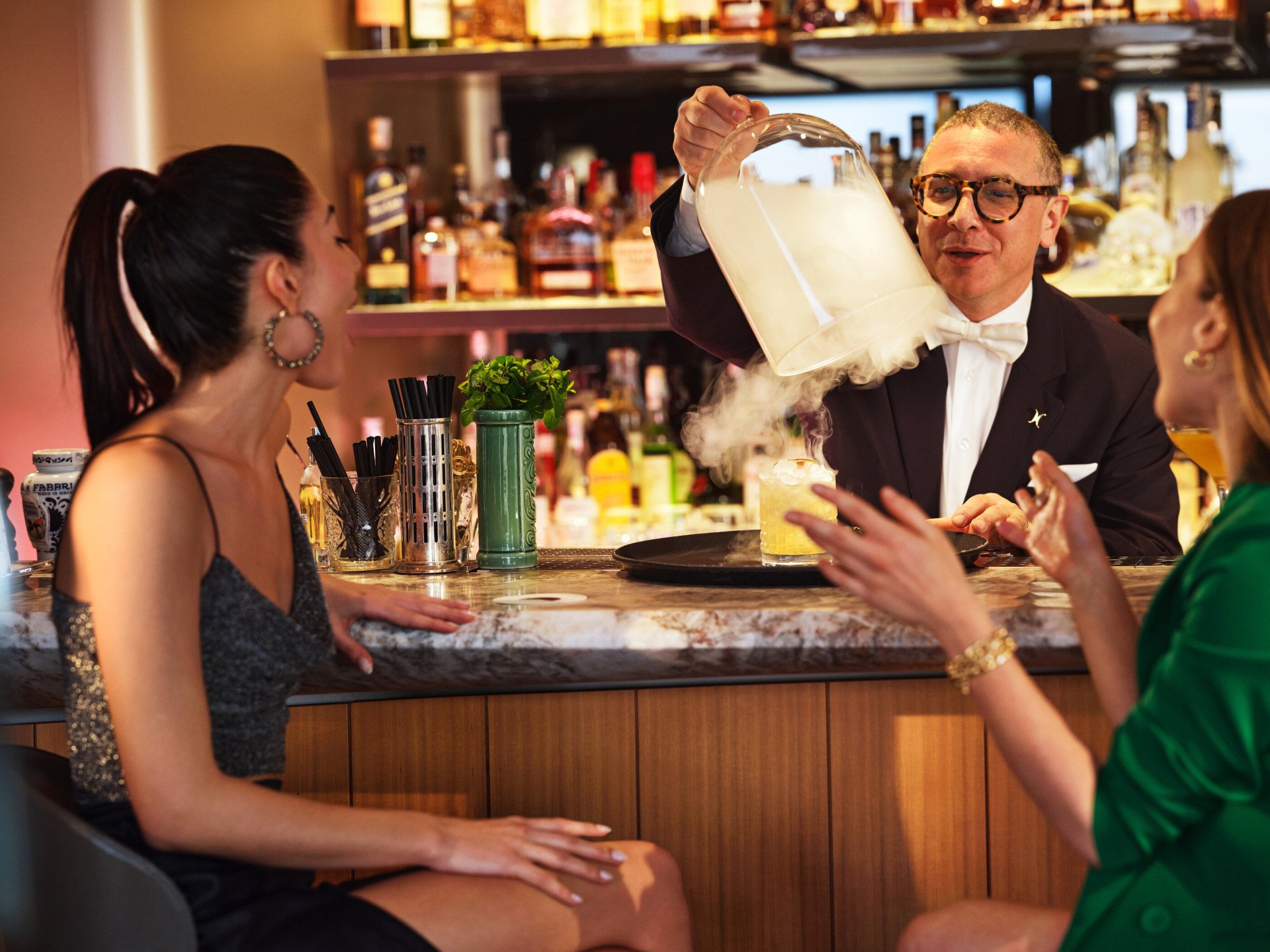 Barman che solleva una campana di vetro e scopre un cocktail per due giovani donne sedute al bancone de Il Giardino Bar