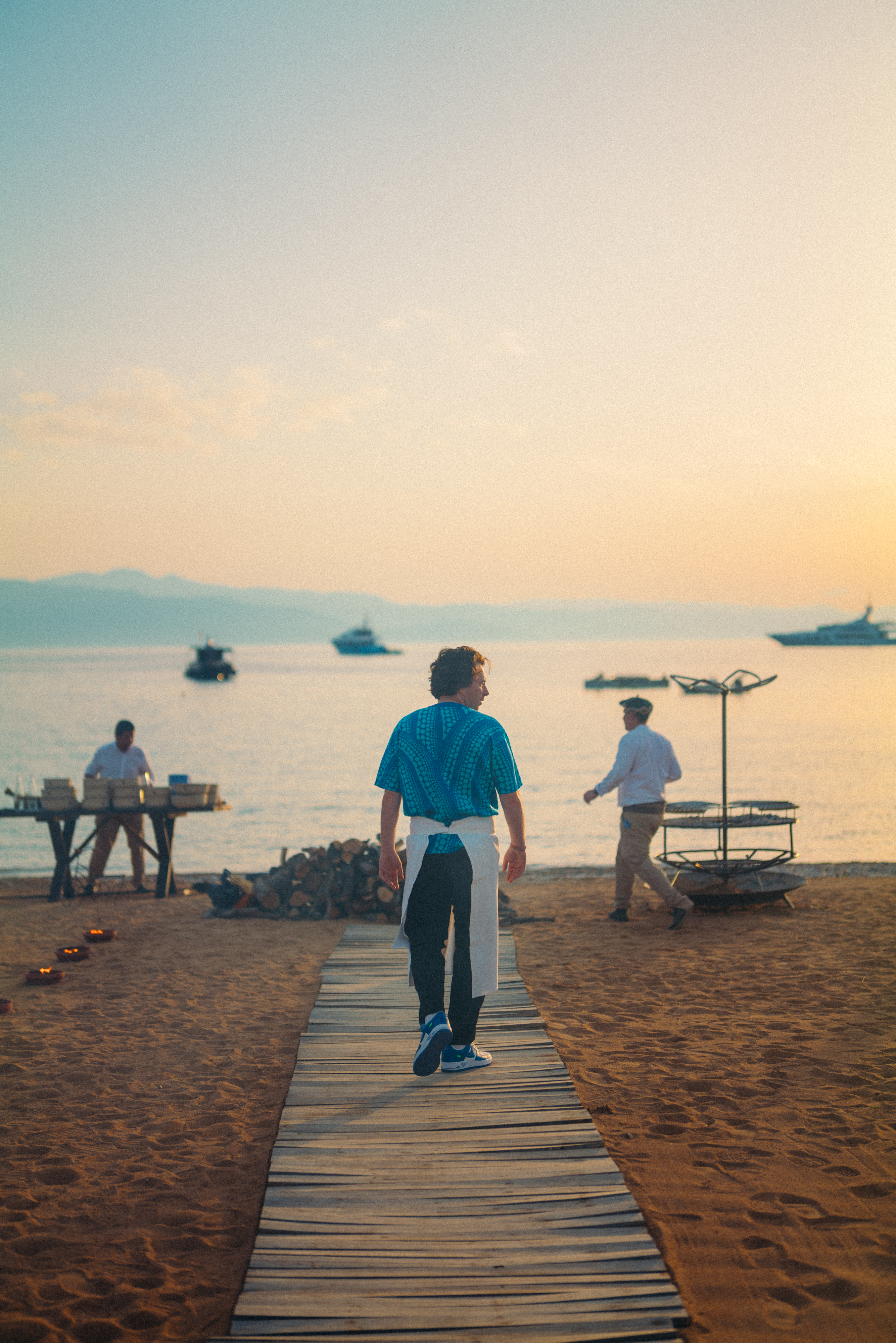 Une personne vêtue d'une chemise bleue marche sur un chemin en bois sur une plage de sable au coucher du soleil, avec des bateaux sur l'eau et des gens qui installent des tables à proximité. Atmosphère paisible.