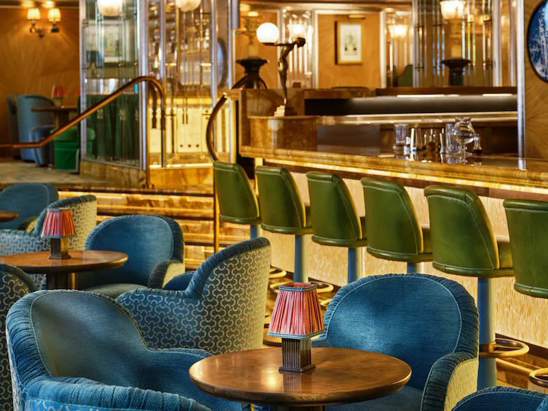 Blue upholstered armchairs and green bar stools at The Vesper Bar, beneath ornate chandeliers and warm Art Deco interiors.