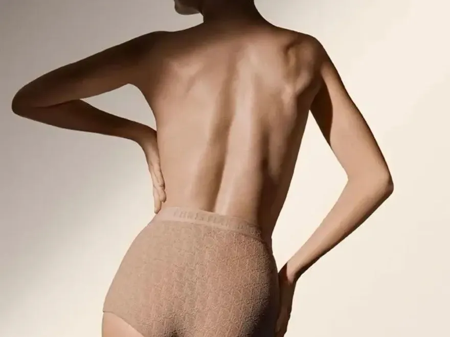 A woman in underwear stands with her hands on her hips, showcasing her figure against an indoor wall.