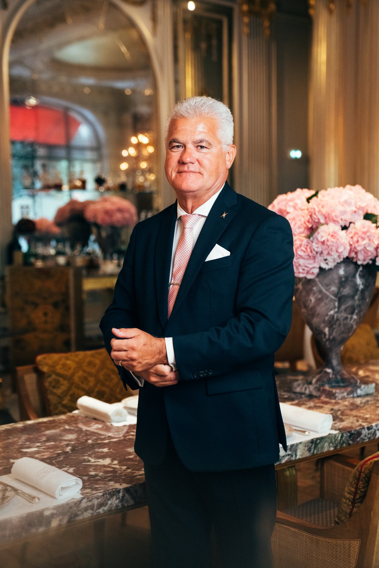 Person in formal suit standing in elegant room with pink flower arrangement and marble table.