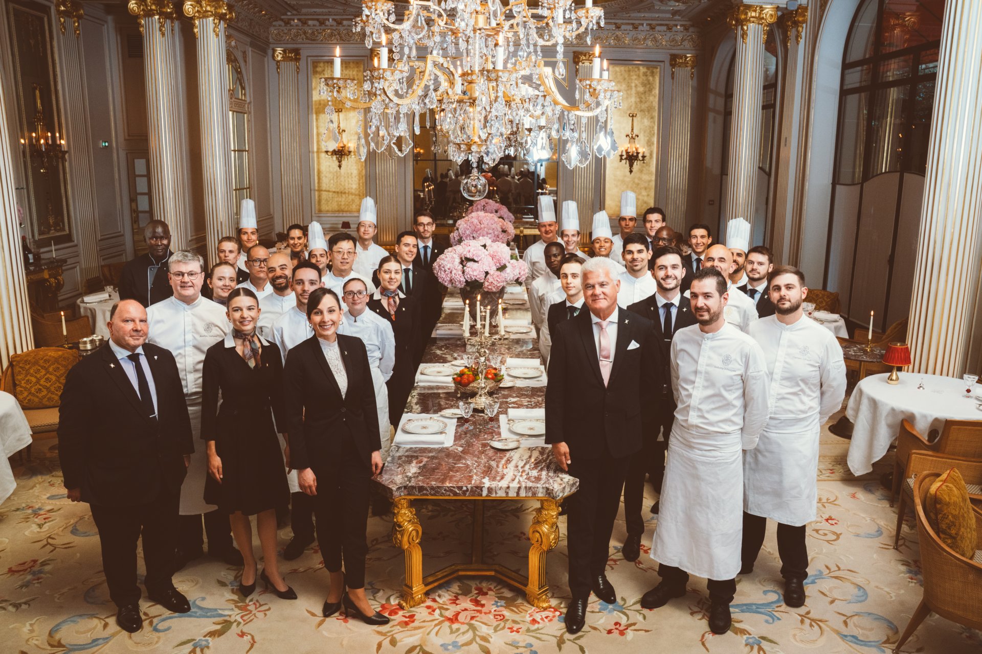Group of people in formal attire and chef uniforms in a decorated dining room with a long table.