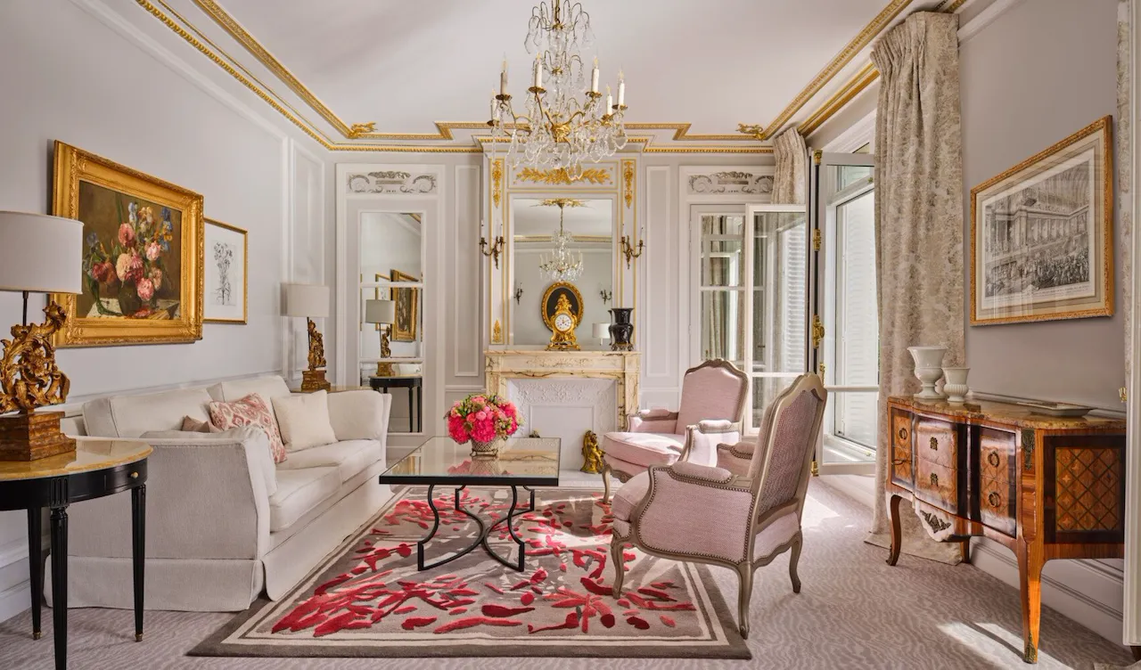 Sala de estar elegante com decoração clássica: sofá branco, poltronas cor-de-rosa, lustre ornamentado, detalhes dourados, arte floral e tapete com padrão vermelho. Ambiente aconchegante e luxuoso.