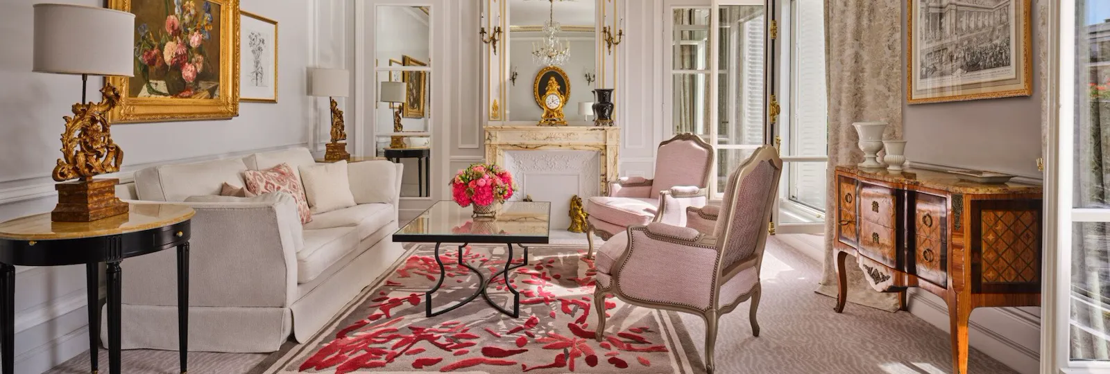 Sala de estar elegante com decoração clássica: sofá branco, poltronas cor-de-rosa, lustre ornamentado, detalhes dourados, arte floral e tapete com padrão vermelho. Ambiente aconchegante e luxuoso.