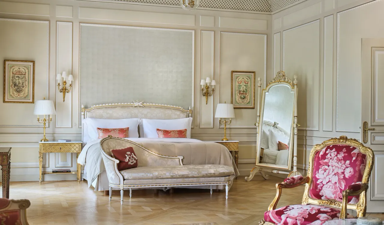 Vista ampla de uma Suíte Versalhes com a cadeira e a mesa em primeiro plano e o sofá, a cama e o espelho ao fundo, no Le Meurice, Paris
