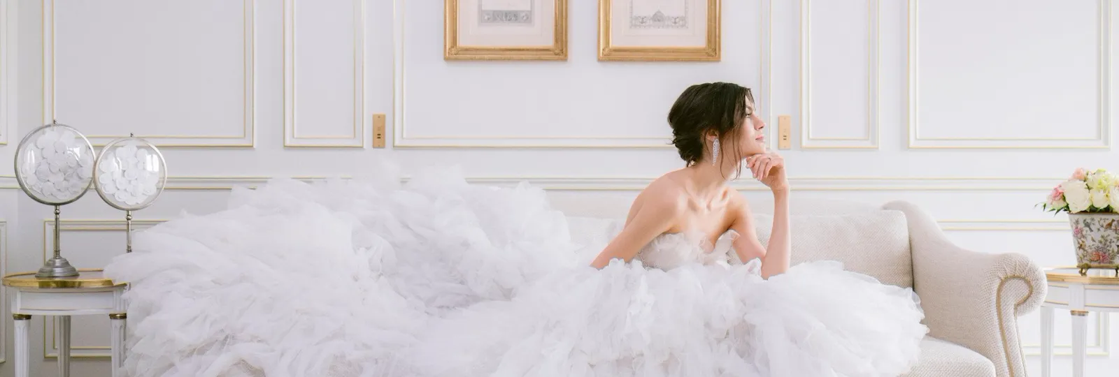 Bride in a large white dress with delicate fabrics, sitting in a suite on a couch and covering it with her dress while looking outside the window with a dreamy eye. On the wall there are two drawings in golden frames, the couch is light beige and the floor covered with a grey/silver printed carpet, at Le Meurice, Paris.