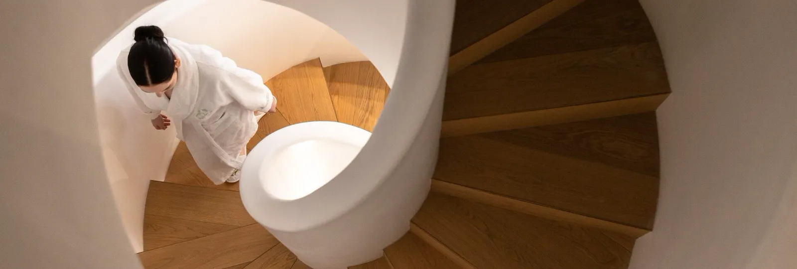 The photo shows a white spiral staircase. The steps are made of wood. A woman in a white bathrobe climbs the stairs. We watch her from above, her black hair tied back in a bun at Le Meurice, Paris.