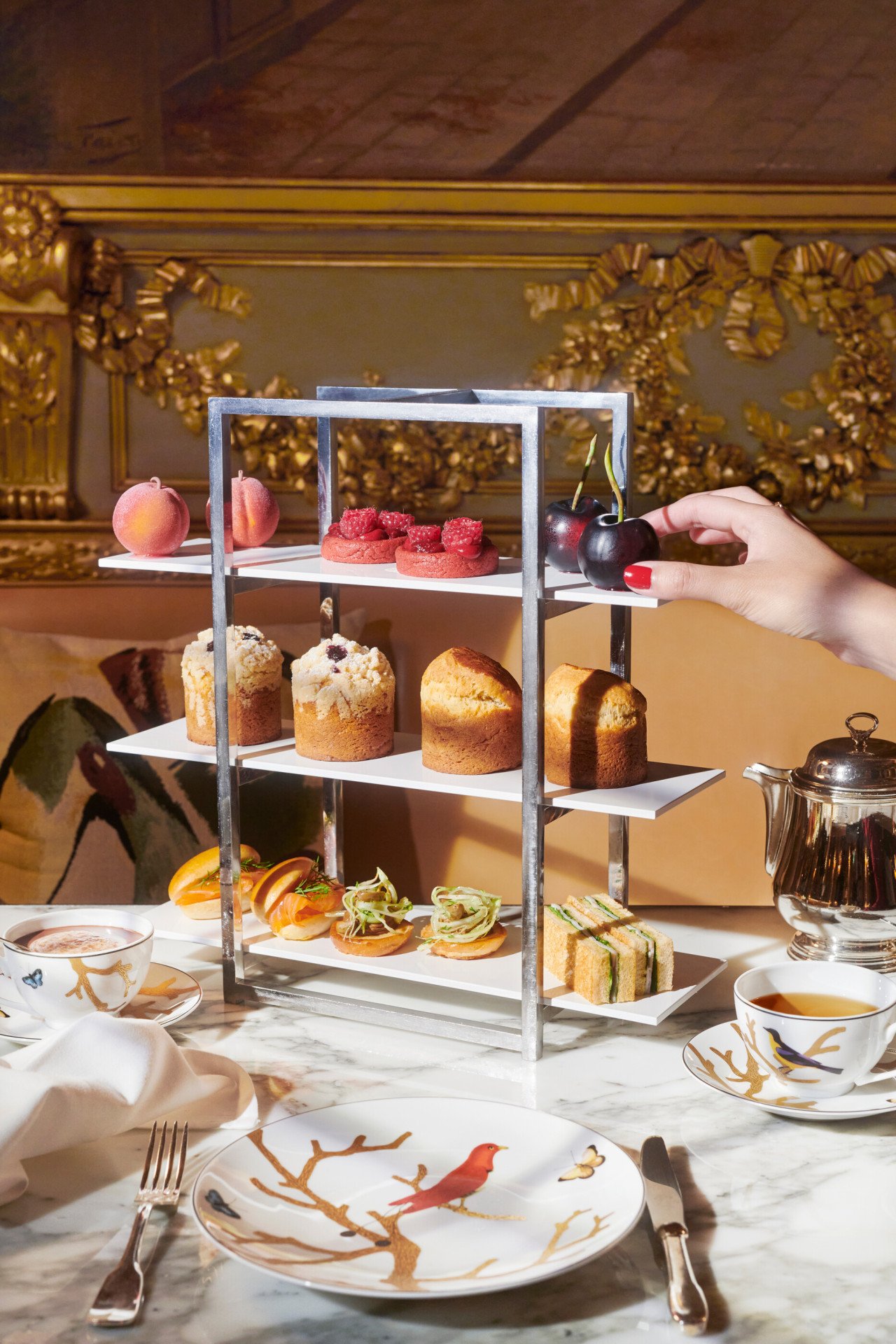 In the foreground, a plate with cultery and two cups of coffee and in the center, a display of cakes with brioche, snaks and fruit by Cédric Grolet that brings a lot of color to the photo, at Le Meurice, Paris