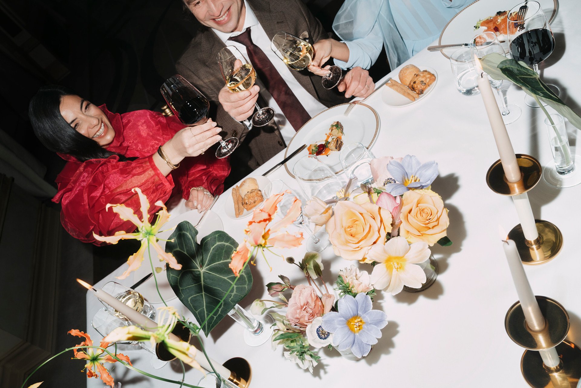 Detail of an event dinner with people eating and drinking red wine, with flowers detail in the foreground 