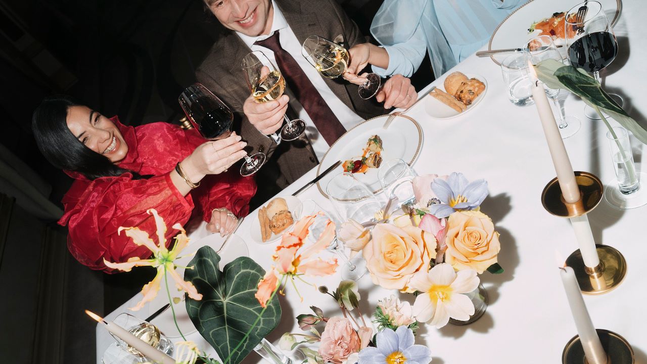 Detail of an event dinner with people eating and drinking red wine, with flowers detail in the foreground
