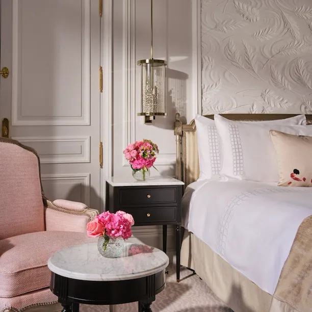 Um quarto luxuoso com uma poltrona rosa macia, uma pequena mesa com tampo de mármore e flores rosa, uma cama com lençóis brancos impecáveis e molduras elegantes nas paredes.