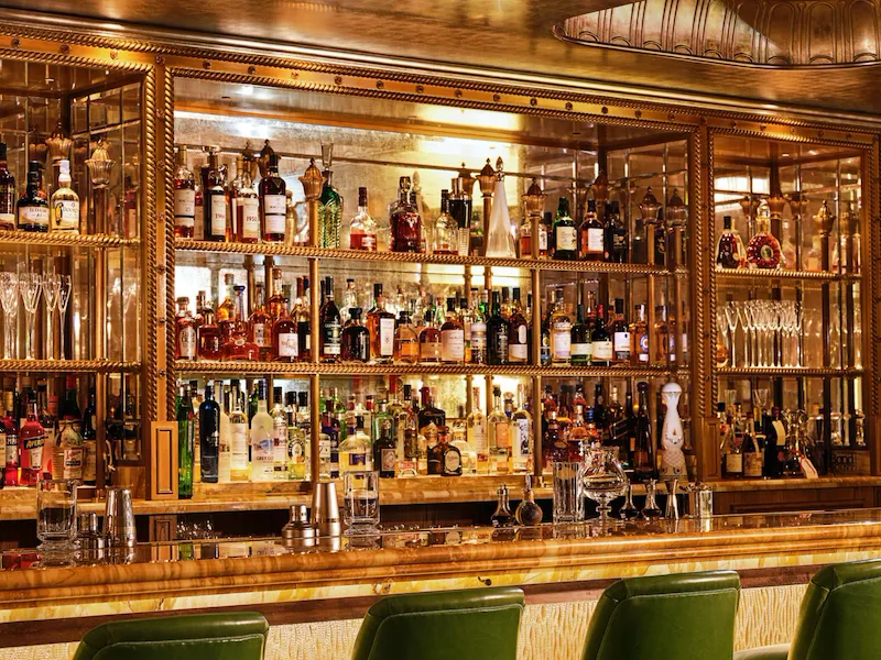 Green leather bar stools line the marble bar at The Vesper Bar, with illuminated shelves displaying an extensive collection of spirits.