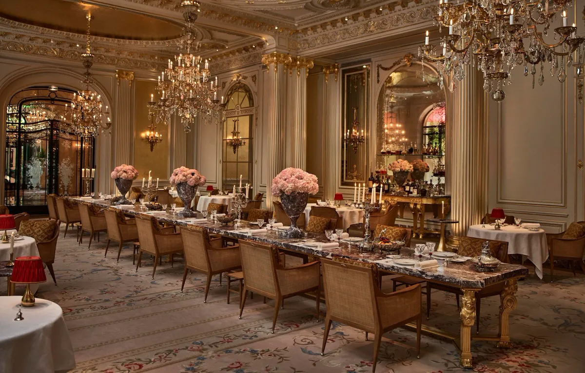 Overview of the Jean Imbert au Plaza Athénée Dining Room and its central marble table with flowers and crystal chandeliers hanging from the ceiling. At Hotel Plaza Athénée, Paris