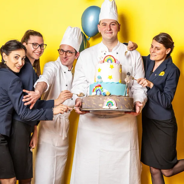 Photo of five employees against a yellow background, one holding a cake with rainbows