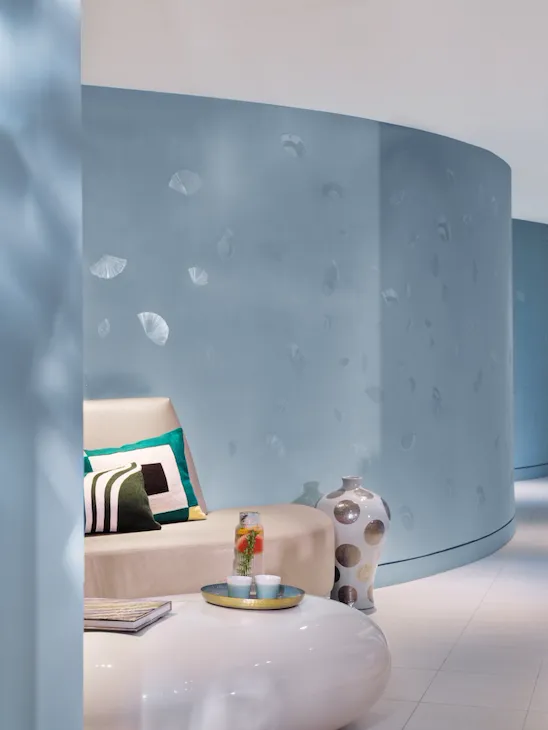 Modern living room with light blue walls, a beige sofa holding geometric cushions, and a large ceramic vase. A glossy coffee table with drinks sits in the foreground.