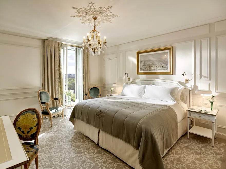 Global view of an executive room highlighting the light grey and cream undertones, as well as the view over the Tuileries gardens in the background. Jewelry and books are on the bedside tablesAt Le Meurice, Paris.