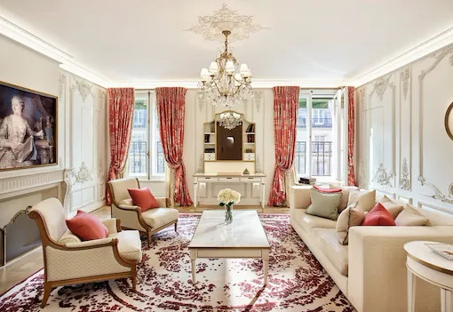 Salon luxueux avec lustre orné, canapés élégants et touches de rouge. Grandes fenêtres avec rideaux rouges, tapis à motifs floraux et tableau classique.