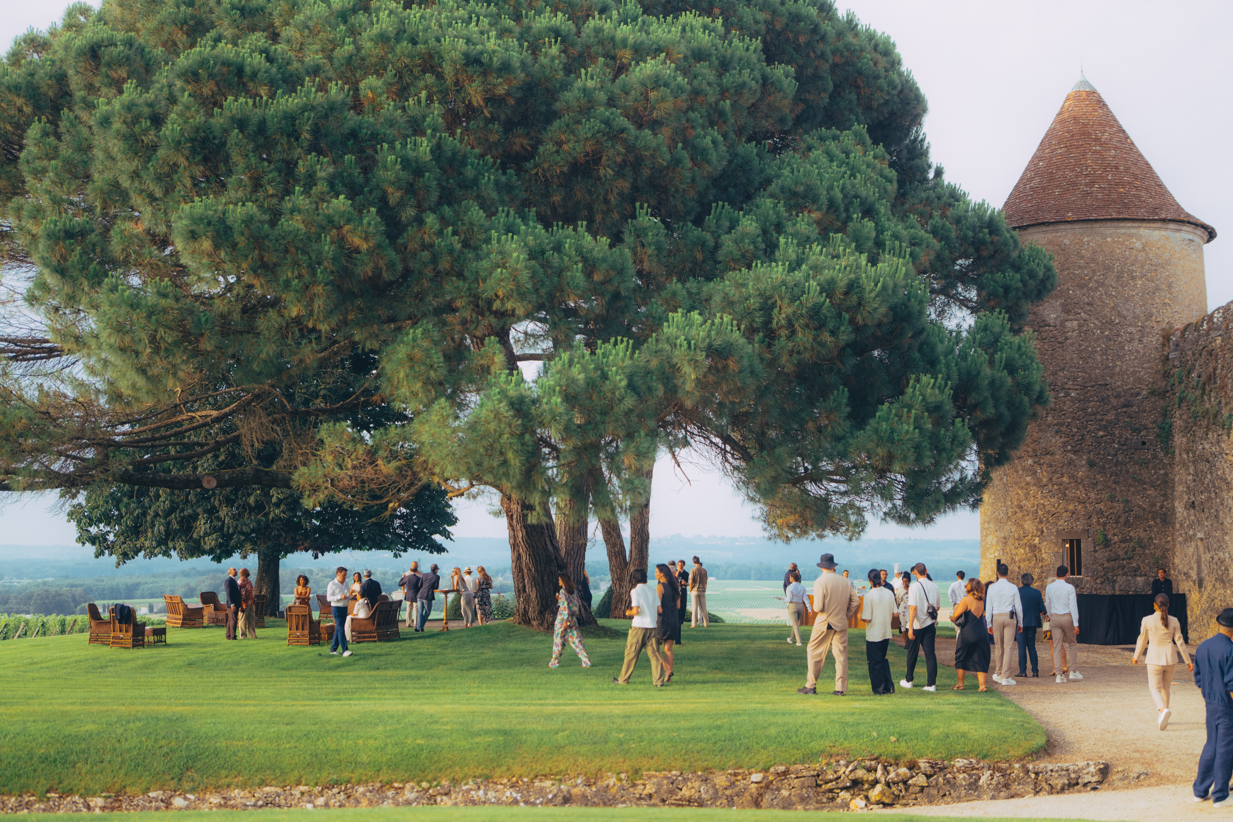 Un groupe de personnes se rassemble sur une pelouse verdoyante, à l'ombre de grands arbres, près d'une ancienne tour en pierre. L'atmosphère est détendue et conviviale, sous un ciel dégagé.