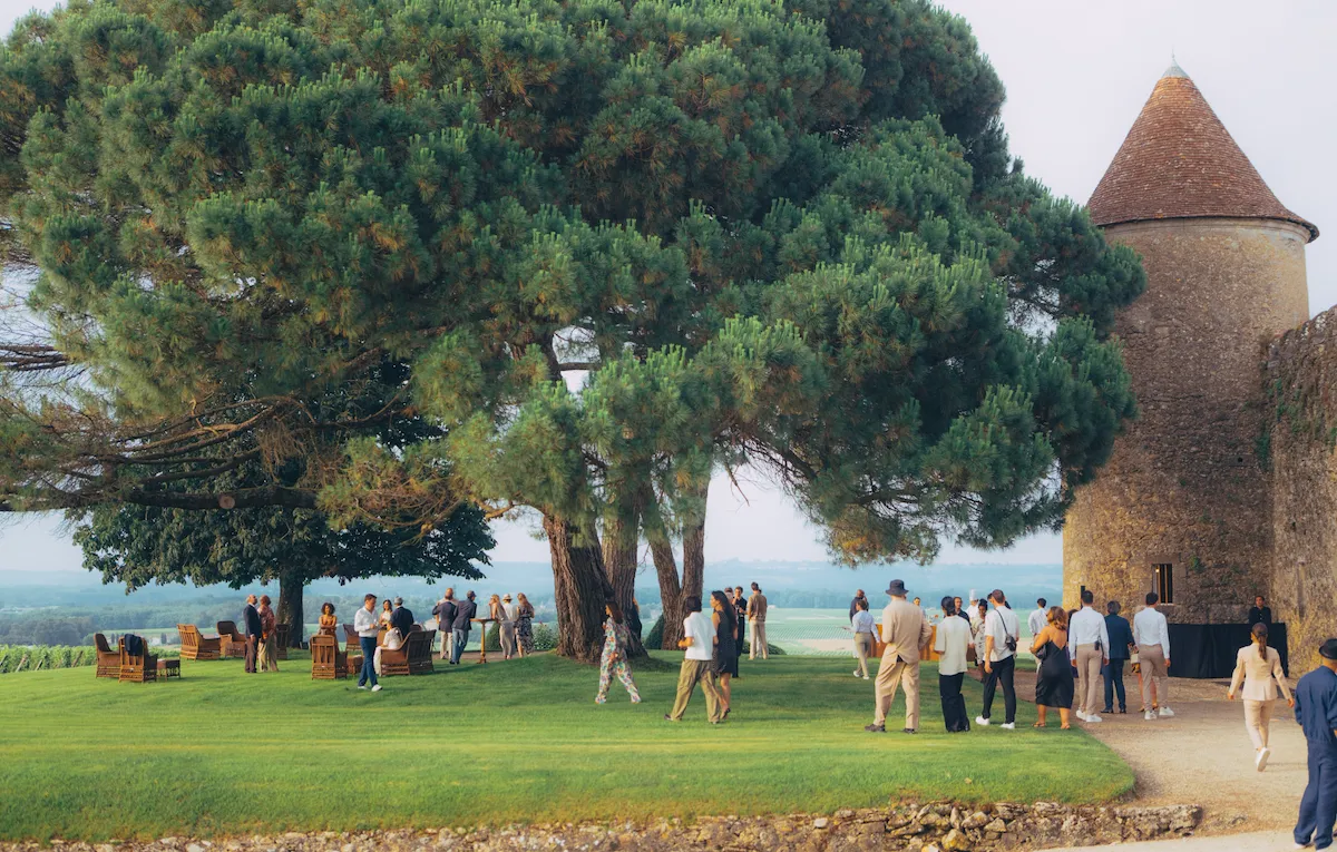 A group of people gather on a lush green lawn under large, shady trees next to an old stone tower. The scene is relaxed and social, with clear skies.