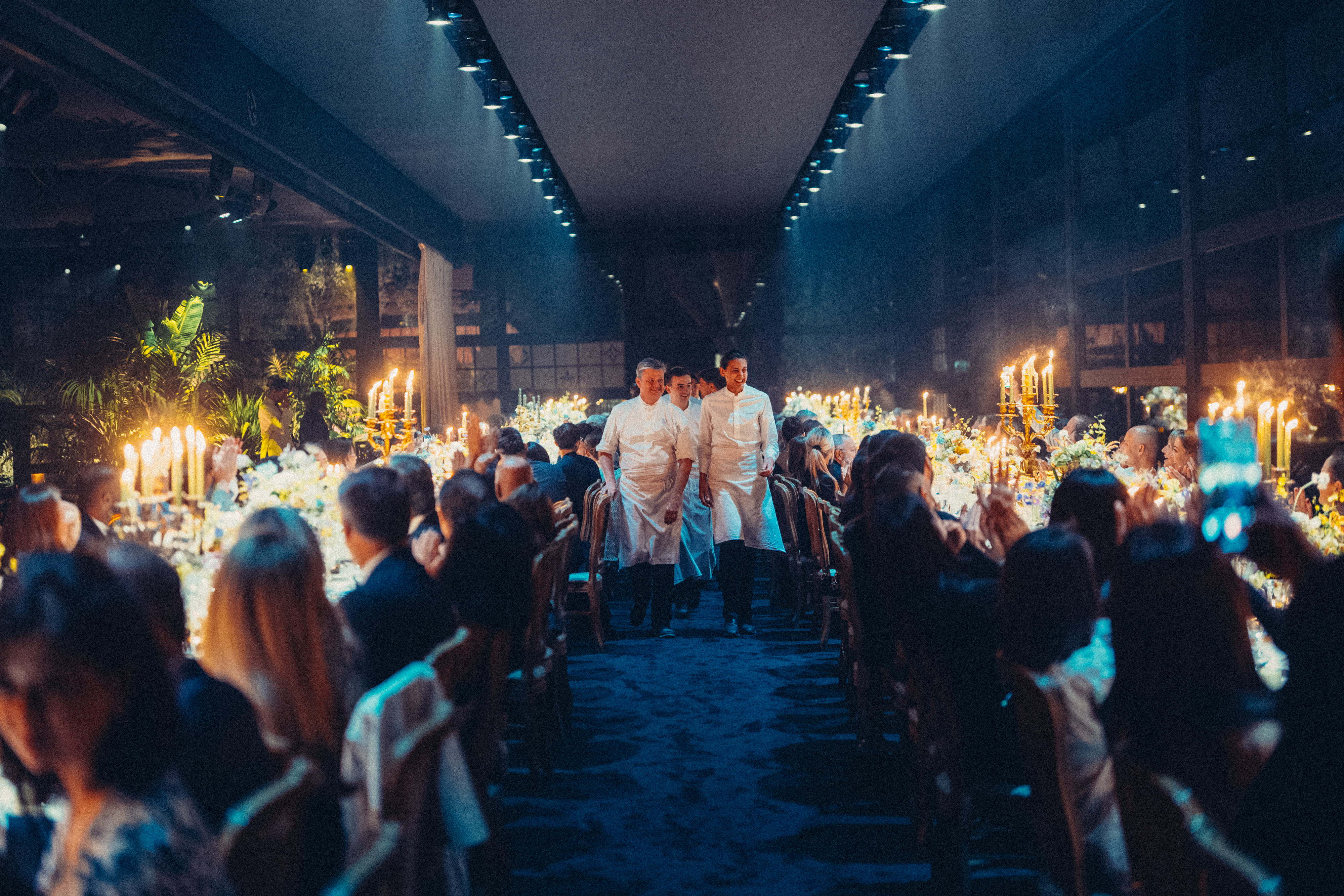 Scène de banquet élégante avec des chefs circulant entre de longues tables ornées de bougies et de fleurs. Les invités sont assis, créant une atmosphère chaleureuse et festive.