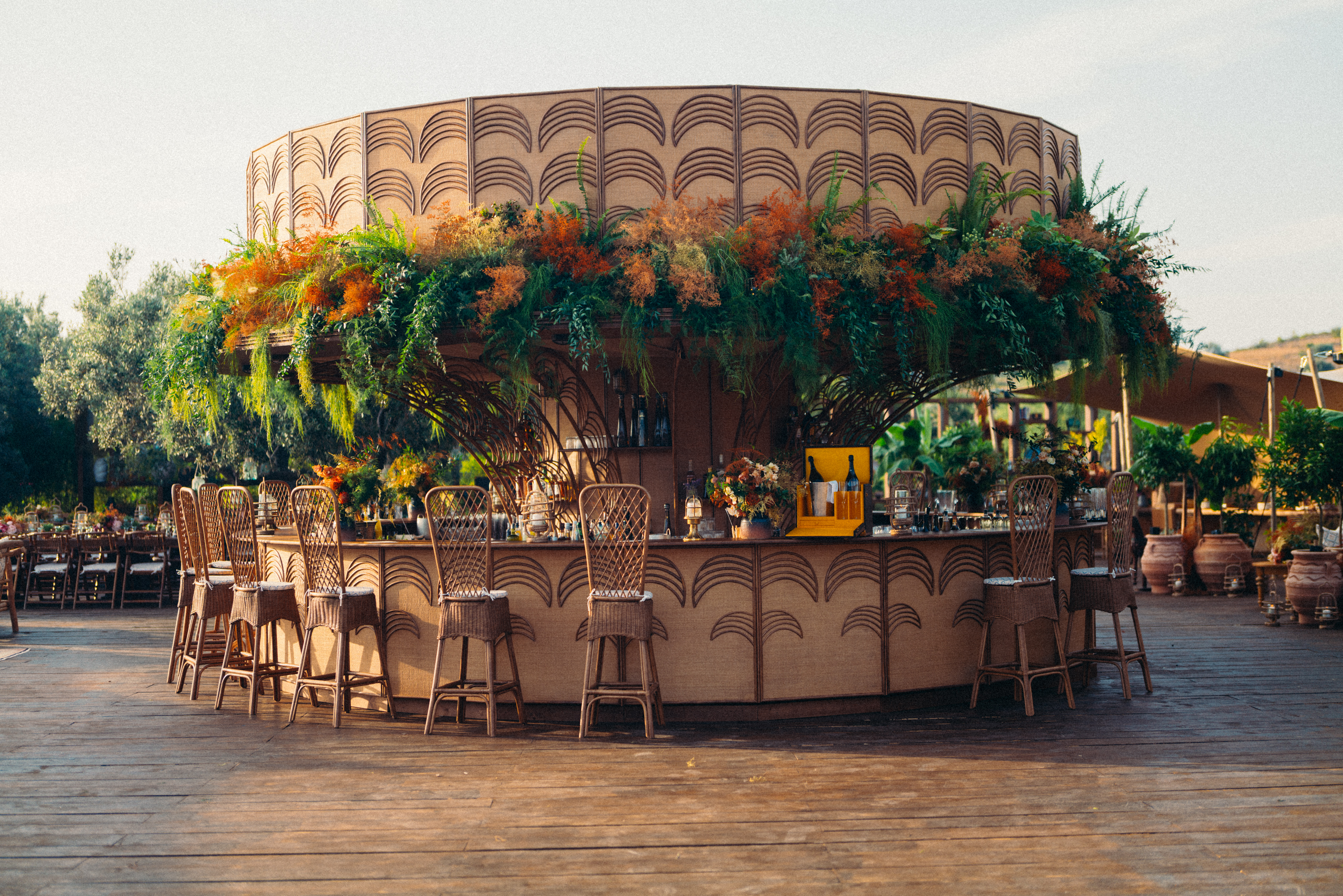 Un bar extérieur rond est agrémenté de hautes chaises en osier et d'une végétation luxuriante aux couleurs orange et verte. Il se trouve sur une terrasse en bois au milieu d'un jardin accueillant et ensoleillé.