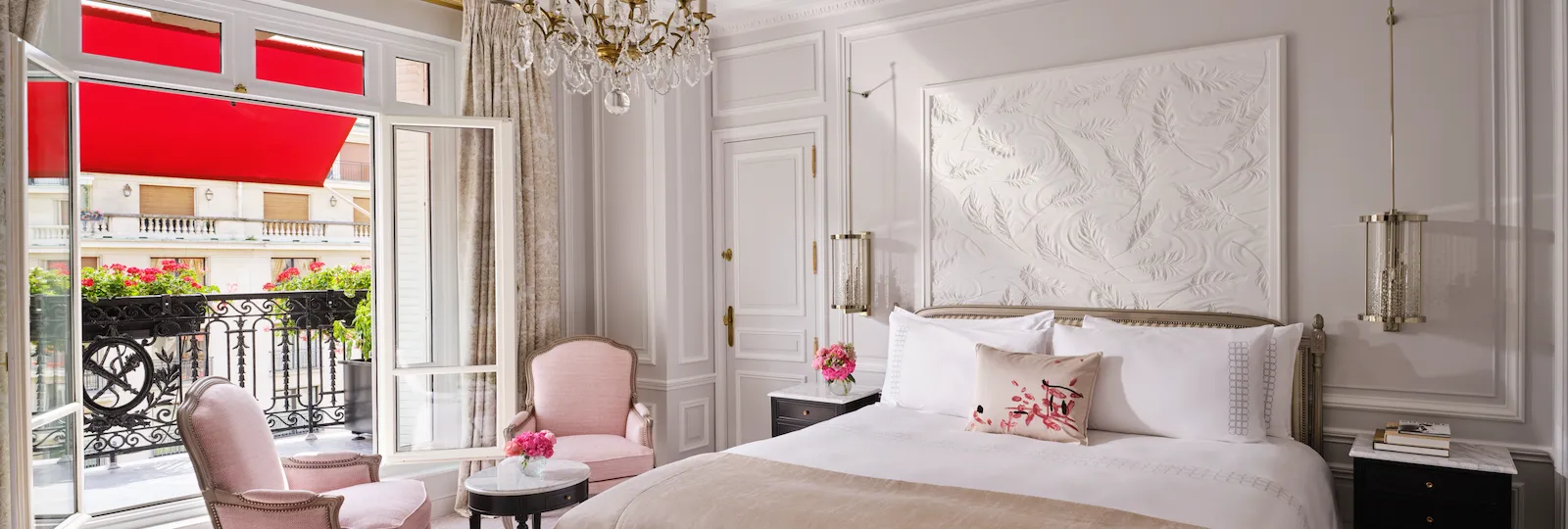 Quarto de hotel elegante com cama luxuosa, lustre ornamentado e poltronas cor-de-rosa. As portas da varanda abertas revelam toldos vermelhos e flores. Ambiente luxuoso.