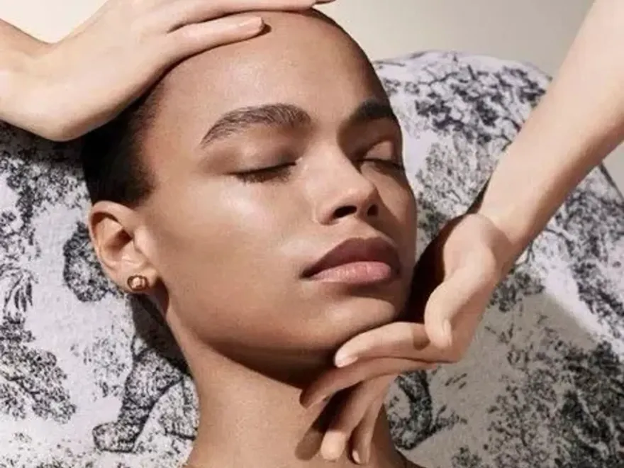 A woman lying down with her eyes closed, showcasing her eyelashes and skin, in a fashion photo shoot setting. The image captures her model features including her shoulder and neck against a wall background.